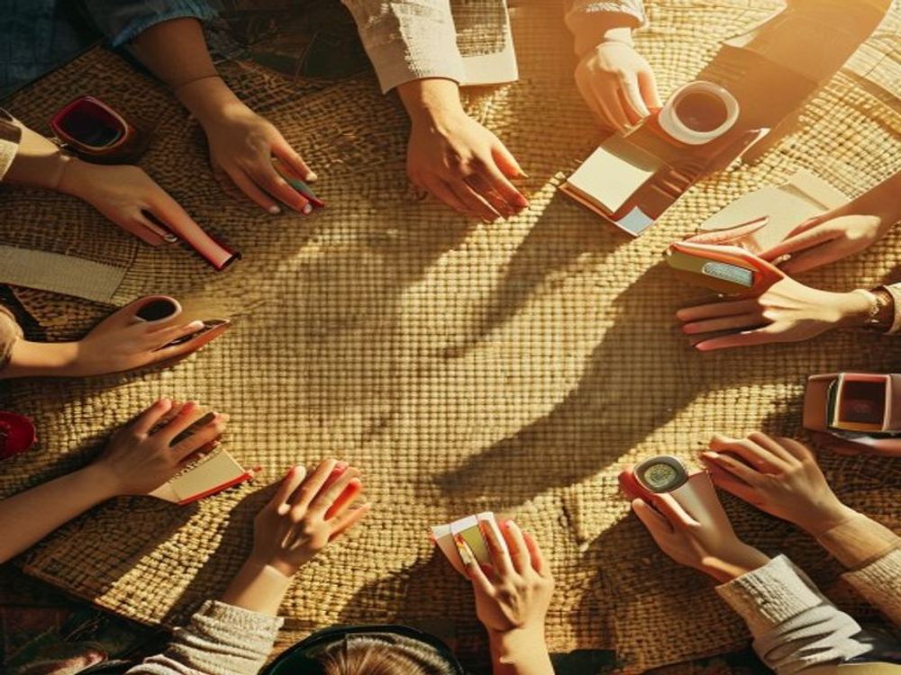 Overhead view of hands around a circle on a woven mat with notebooks and chai cups